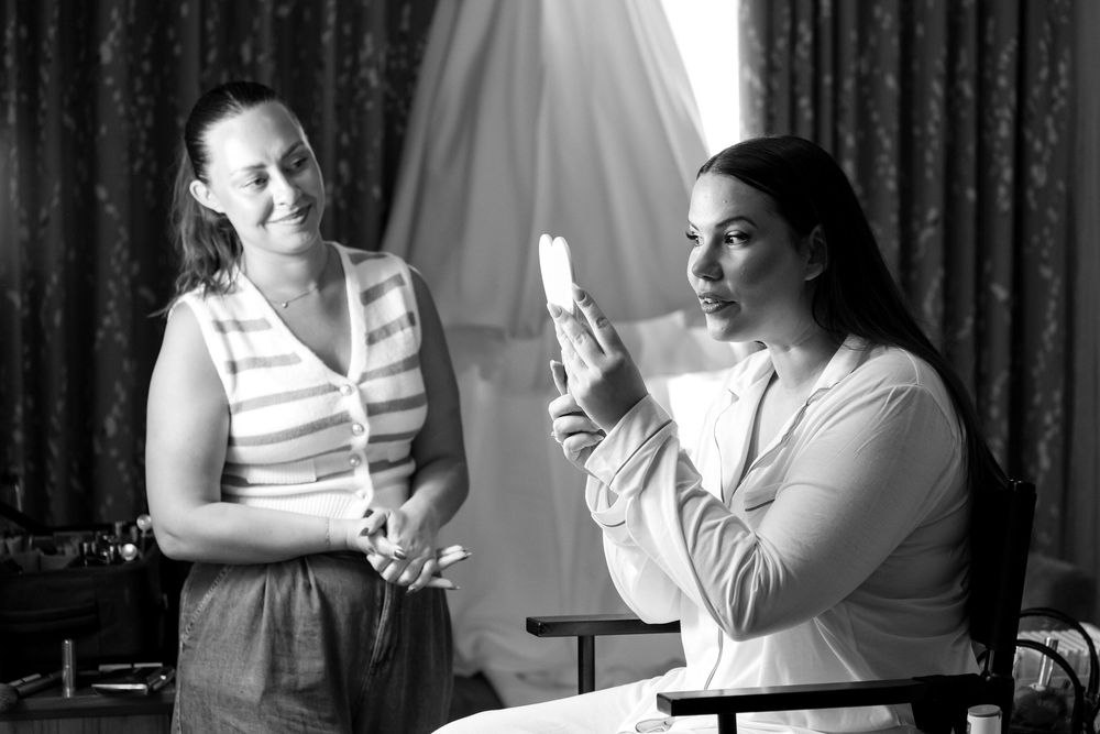 Bride looking into a handheld mirror as her makeup artist looks on during wedding prep.
