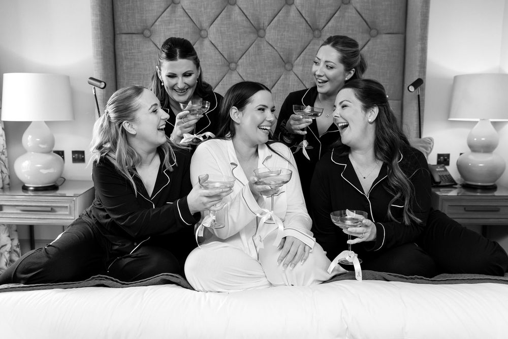 Bride and bridesmaids in matching pyjamas enjoying champagne and laughter on the bed.