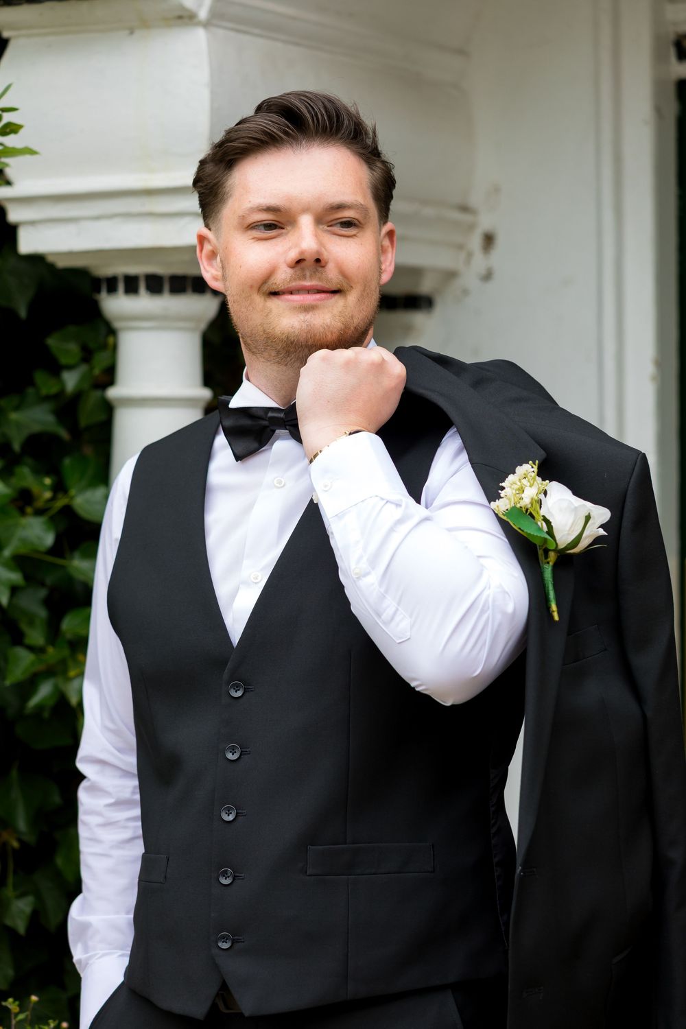 Stylish groom posing confidently with his tuxedo jacket slung over his shoulder.