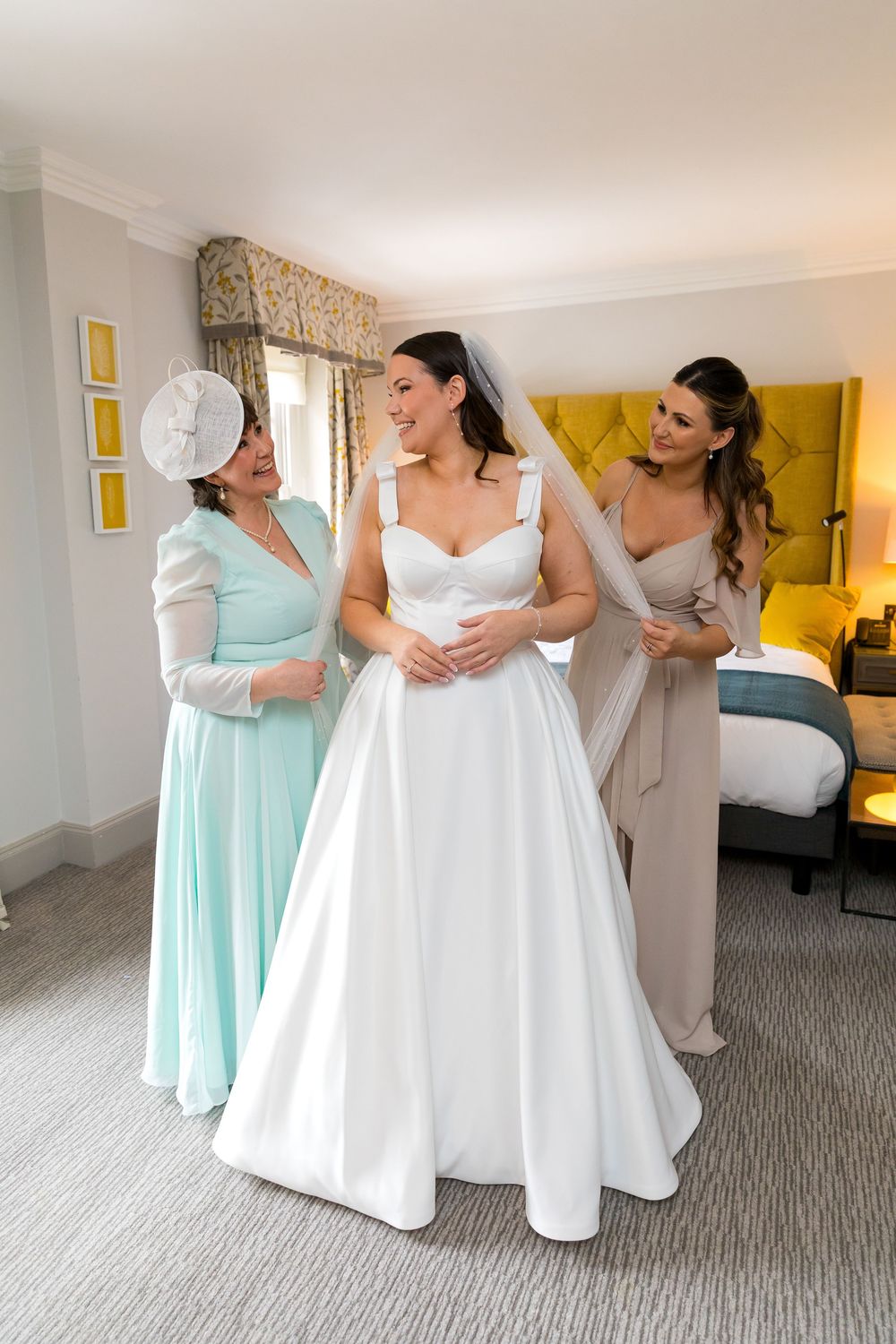 Bride smiling with her mum and bridesmaid helping with the veil in the bridal suite.