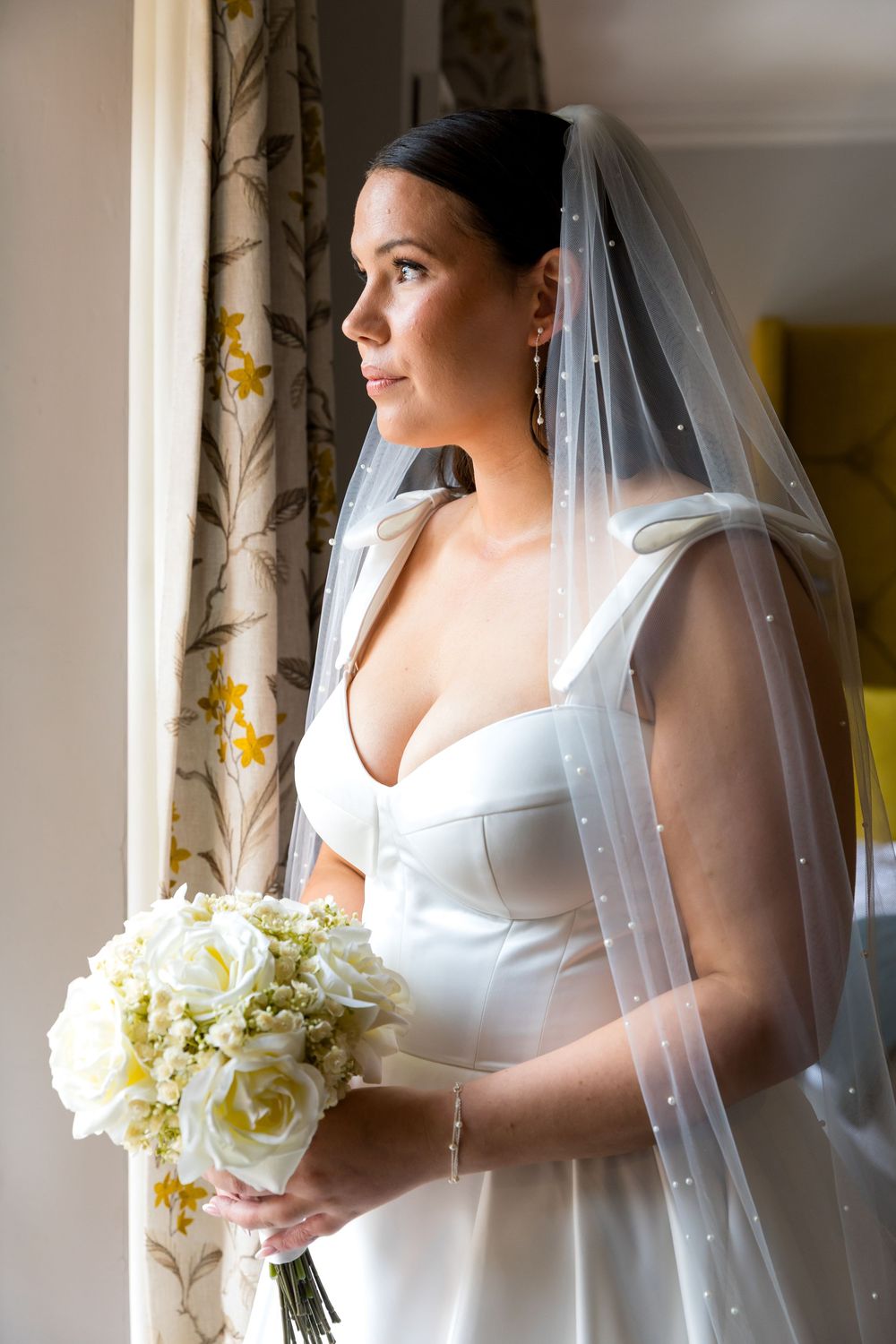 Bride looking out of the window holding a white rose bouquet before the ceremony.