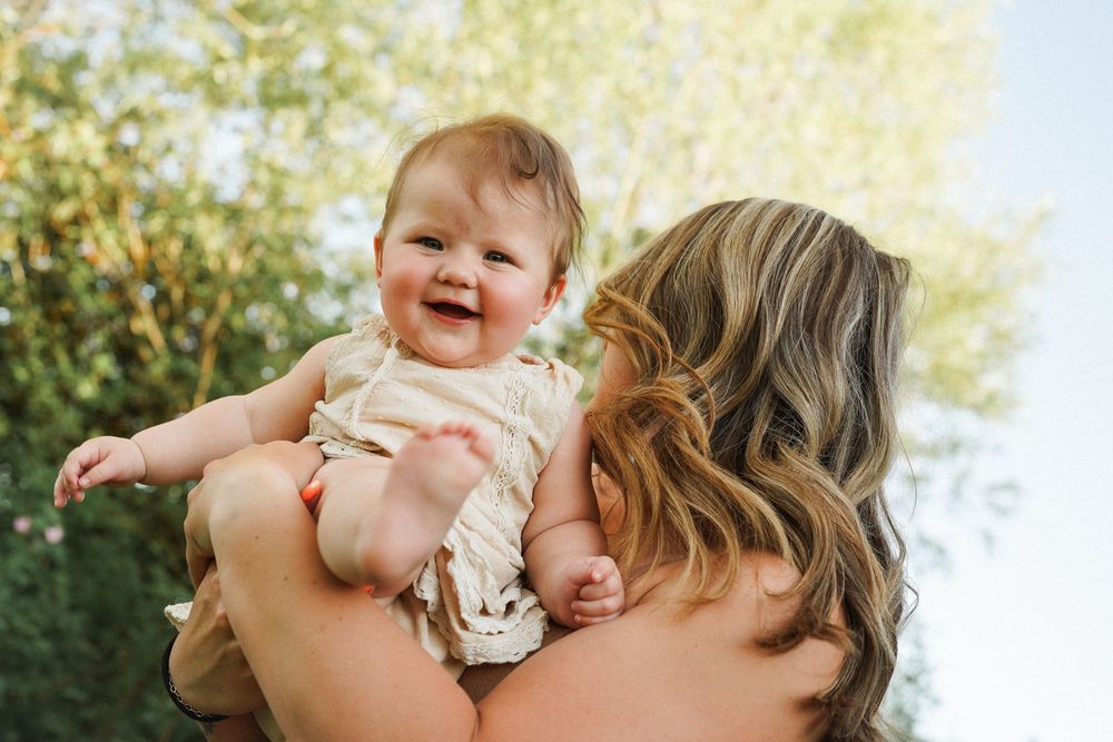 Happy baby wearing light colored outfit being held up outdoors with sunlit foliage in background.
