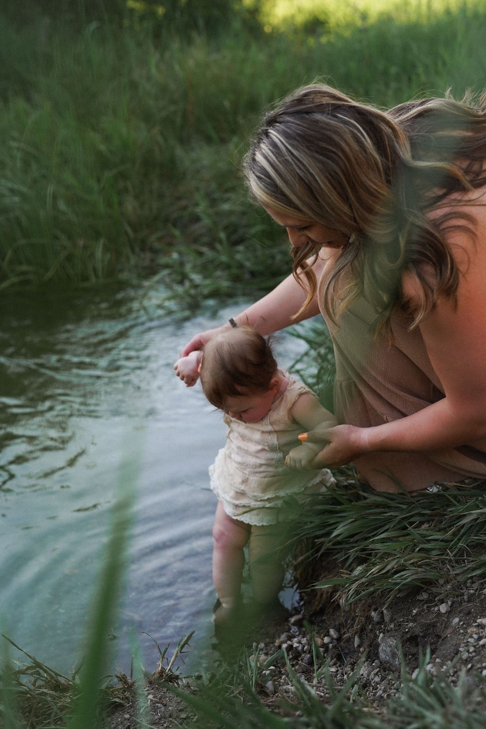 A quiet moment at the water's edge as a parent gently holds a baby near a green shoreline on a summer day.