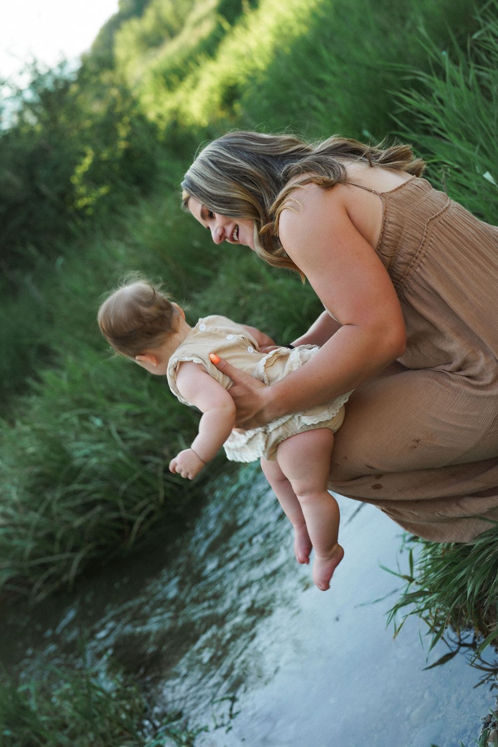 A tender moment by a stream as someone holds a baby near the water's edge on a sunny summer day.