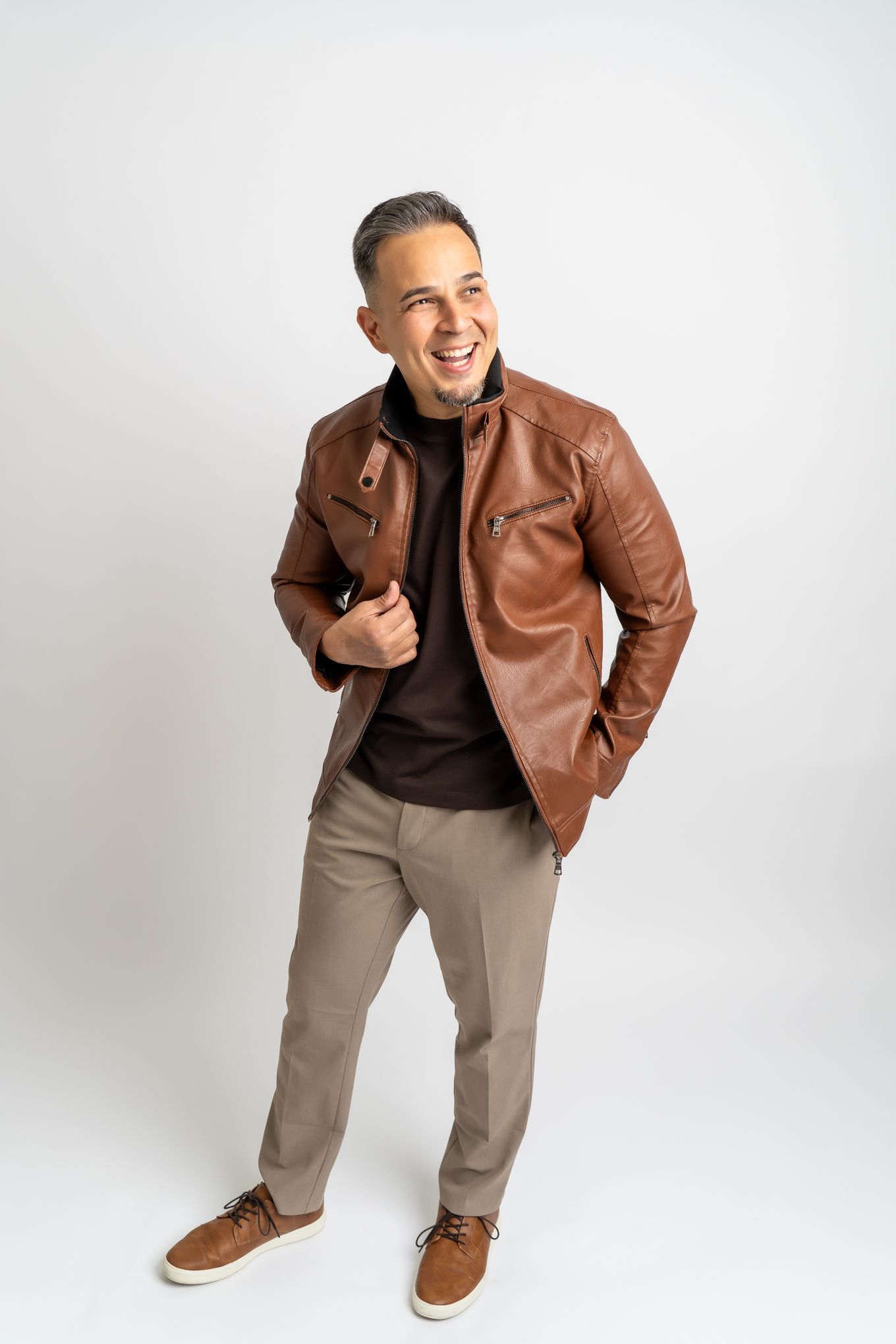 A person wearing a brown leather jacket and khaki pants stands smiling against a white background, striking a casual pose.