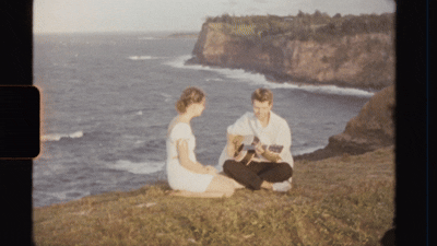 A couple sits on a grassy oceanside cliff while someone plays an acoustic guitar on a sunny day.