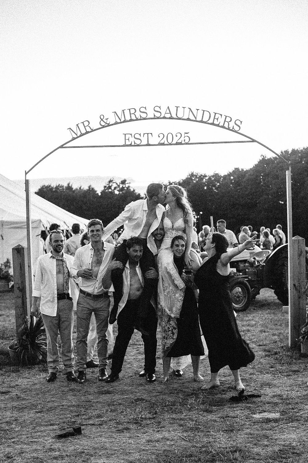 Black and white wedding photo of Mr & Mrs Saunders Est 2025, couple kissing atop guests shoulders under decorative arch.