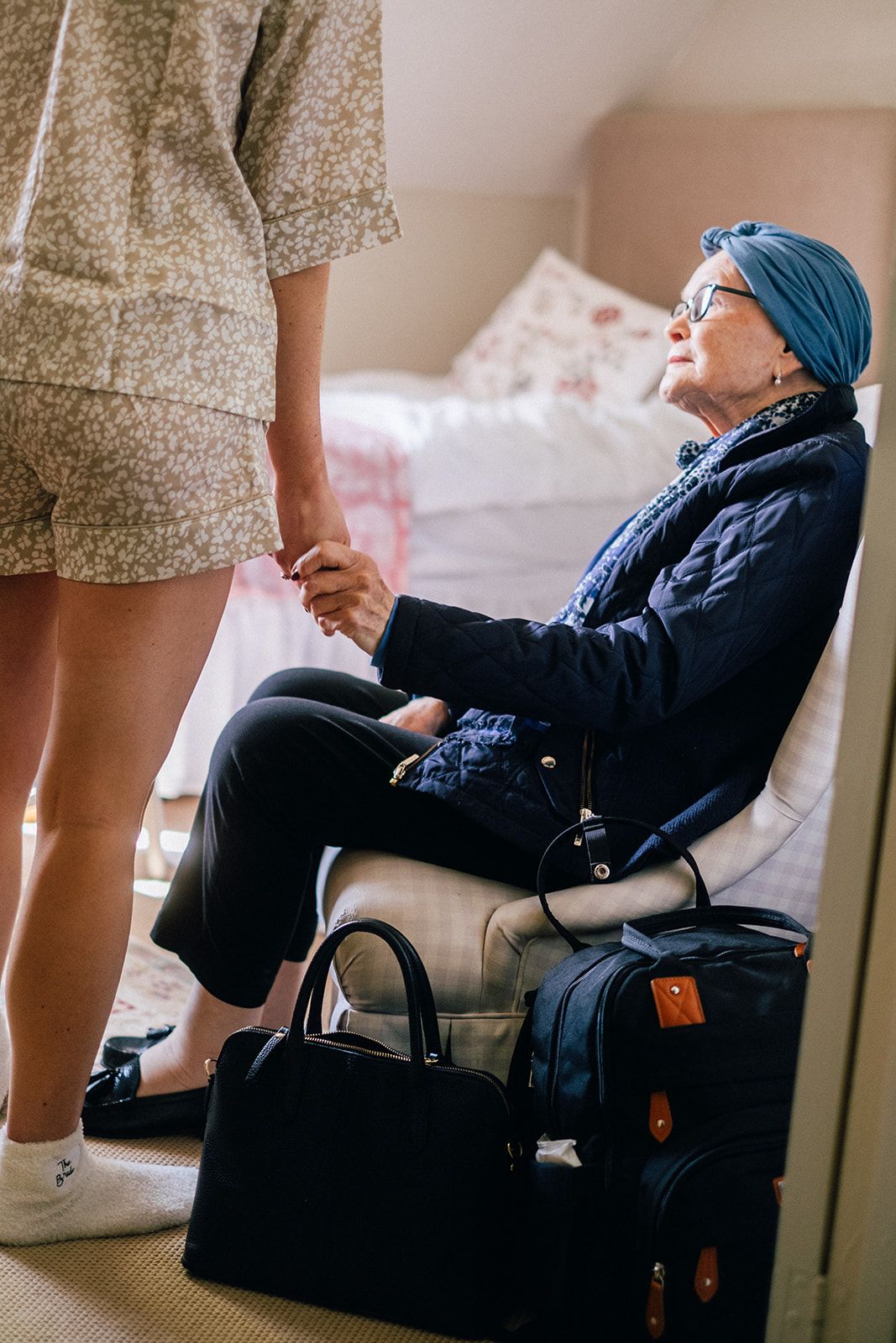 Elderly patient in blue headscarf and glasses holds caregiver's hand while sitting in chair with travel bags nearby.
