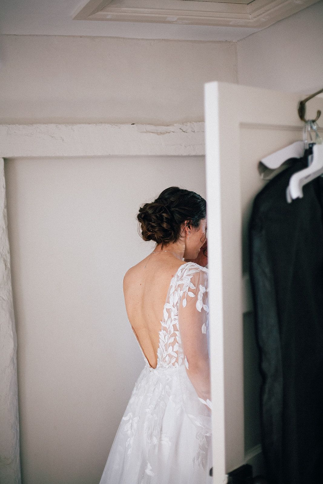 A bridal gown with intricate white lace details and open back design hangs in a minimalist white room.