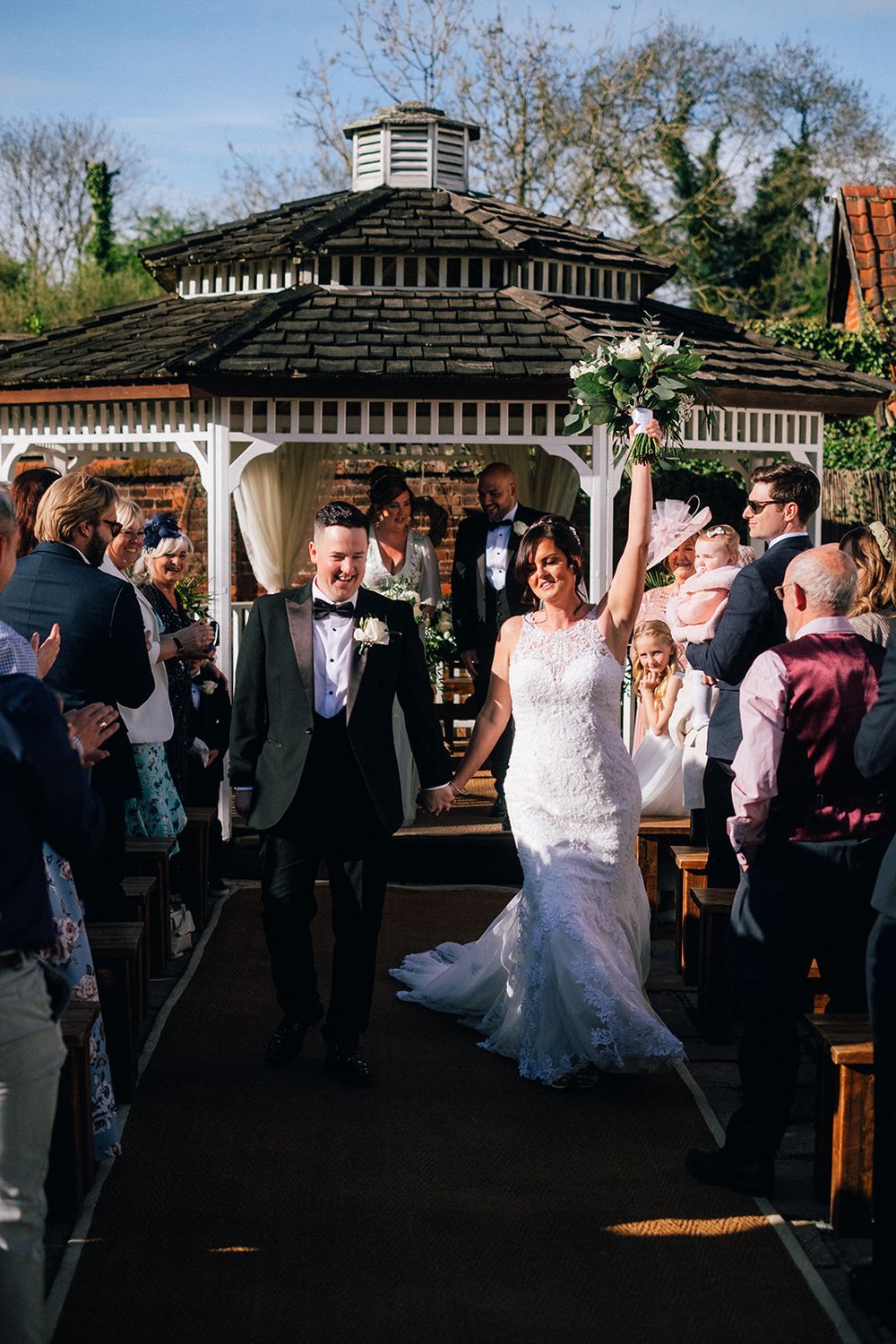 Wedding celebration taking place under a decorative garden gazebo with guests gathered around on a sunny day.