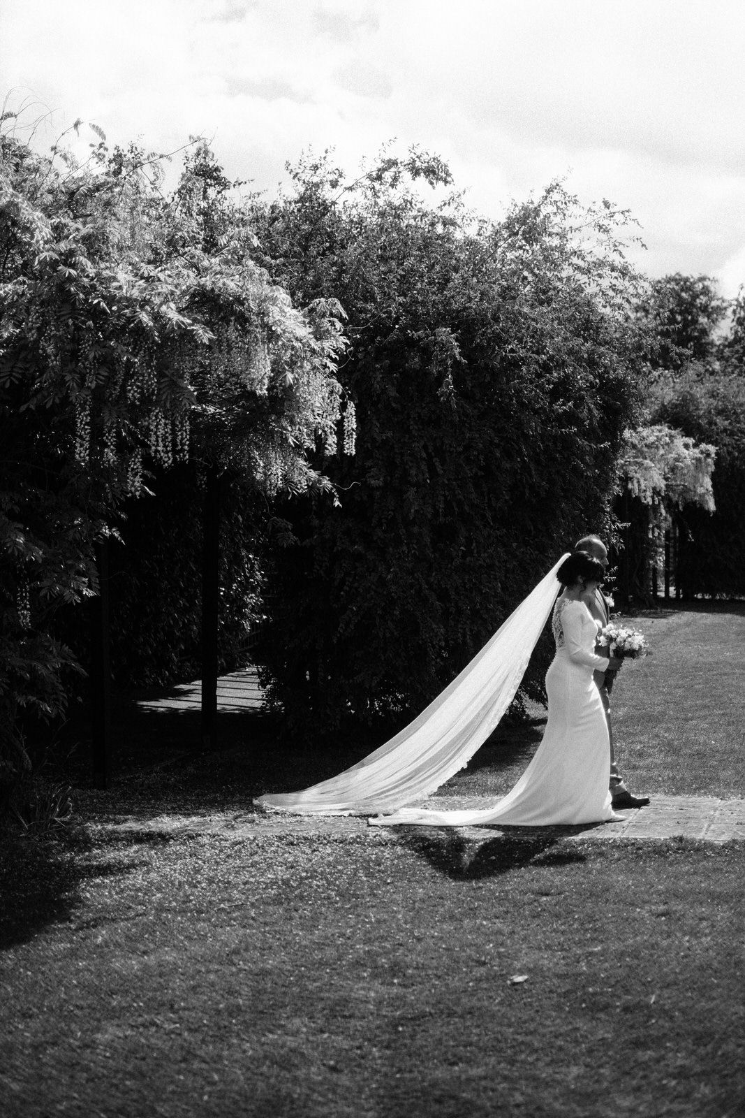 A bride in a flowing white dress and long veil walks across a garden lawn with weeping willow trees in the background.
