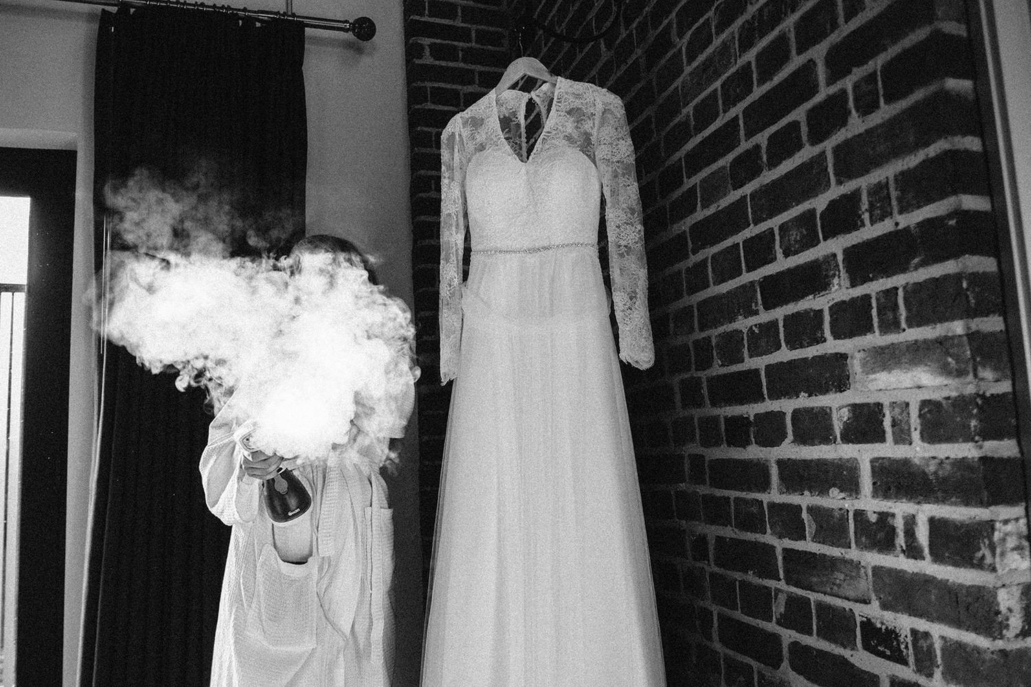 Wedding dress hangs against exposed brick wall with ethereal smoke effect in black and white.