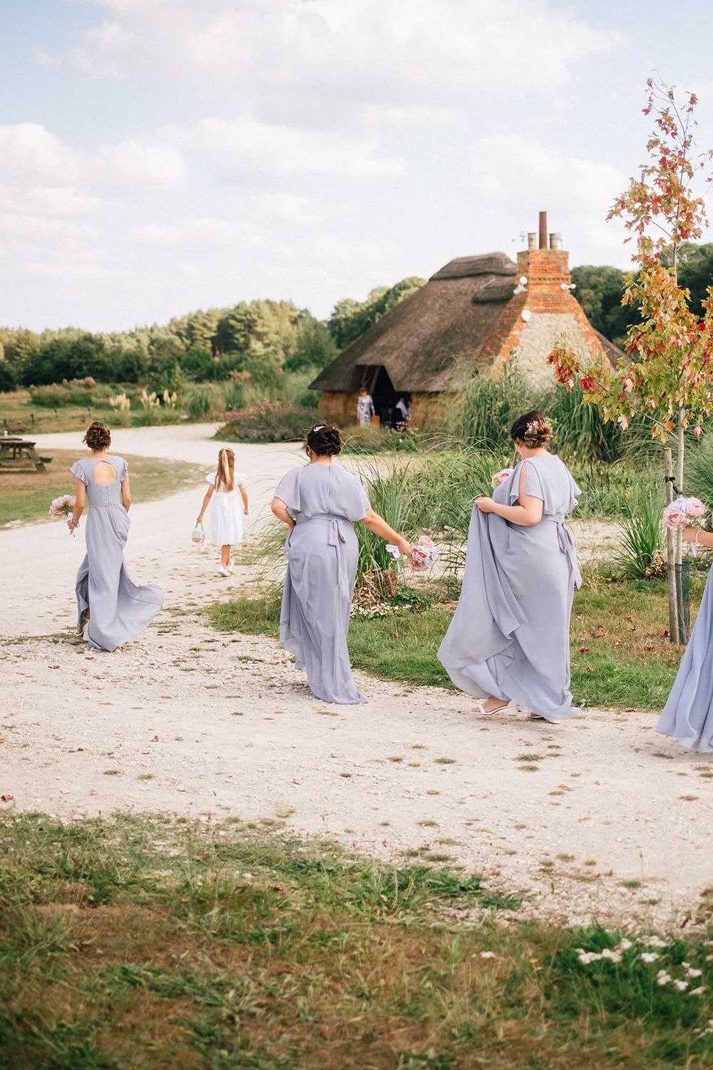 Bridesmaids in soft gray dresses walk along a countryside path near a thatched-roof cottage.