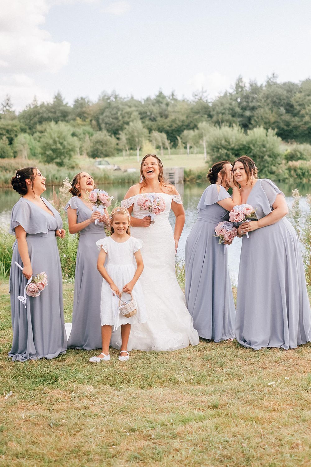 Wedding party in lavender dresses poses for candid laughter-filled photo on grassy lawn.