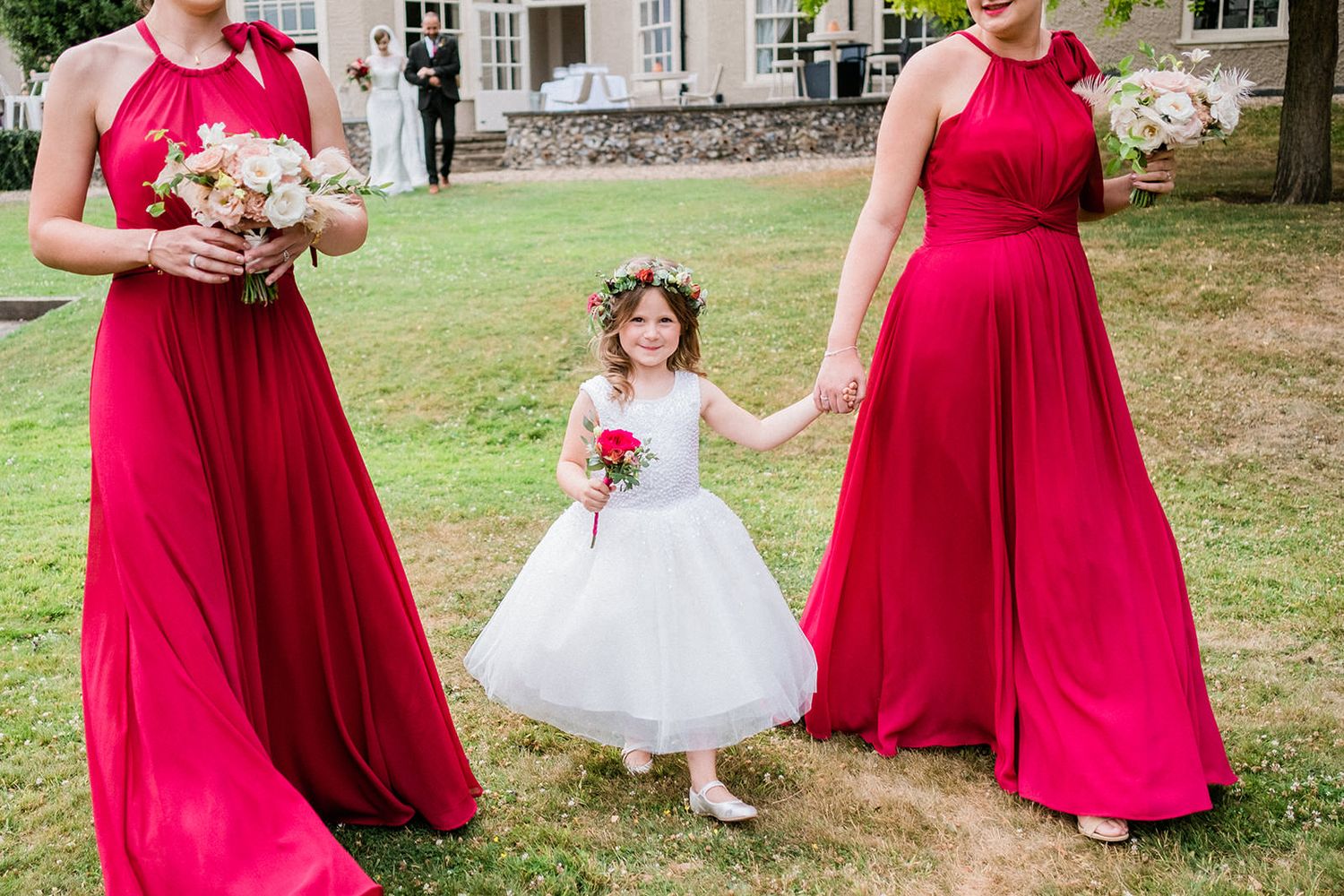 Flower girl in white dress holds hands with bridesmaids wearing vibrant red gowns.