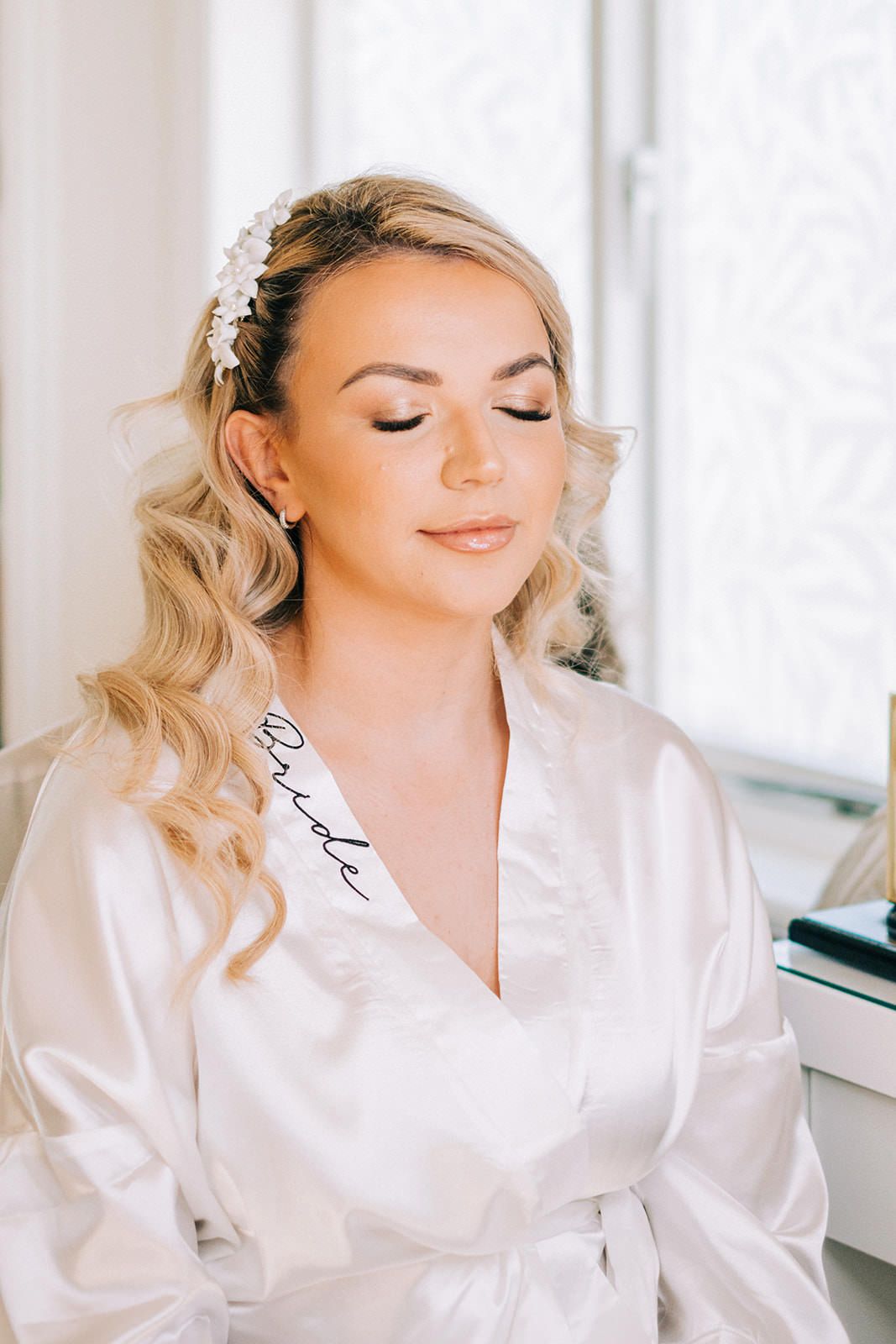 A bride in a white satin robe with blonde curled hair and a crystal hairpiece prepares for a special occasion.