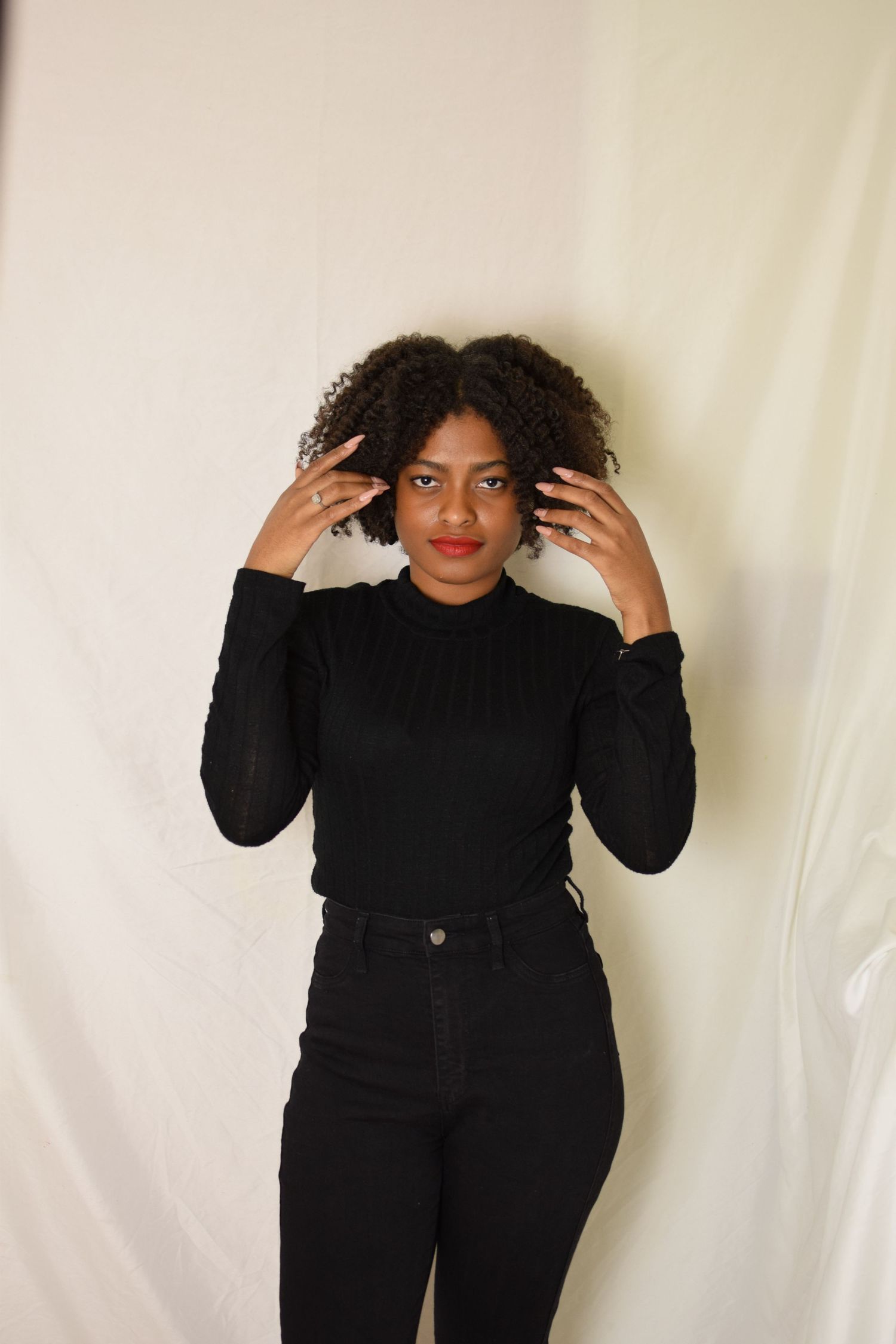 A person in all black clothing poses against a white backdrop with hands raised near their curly hair.