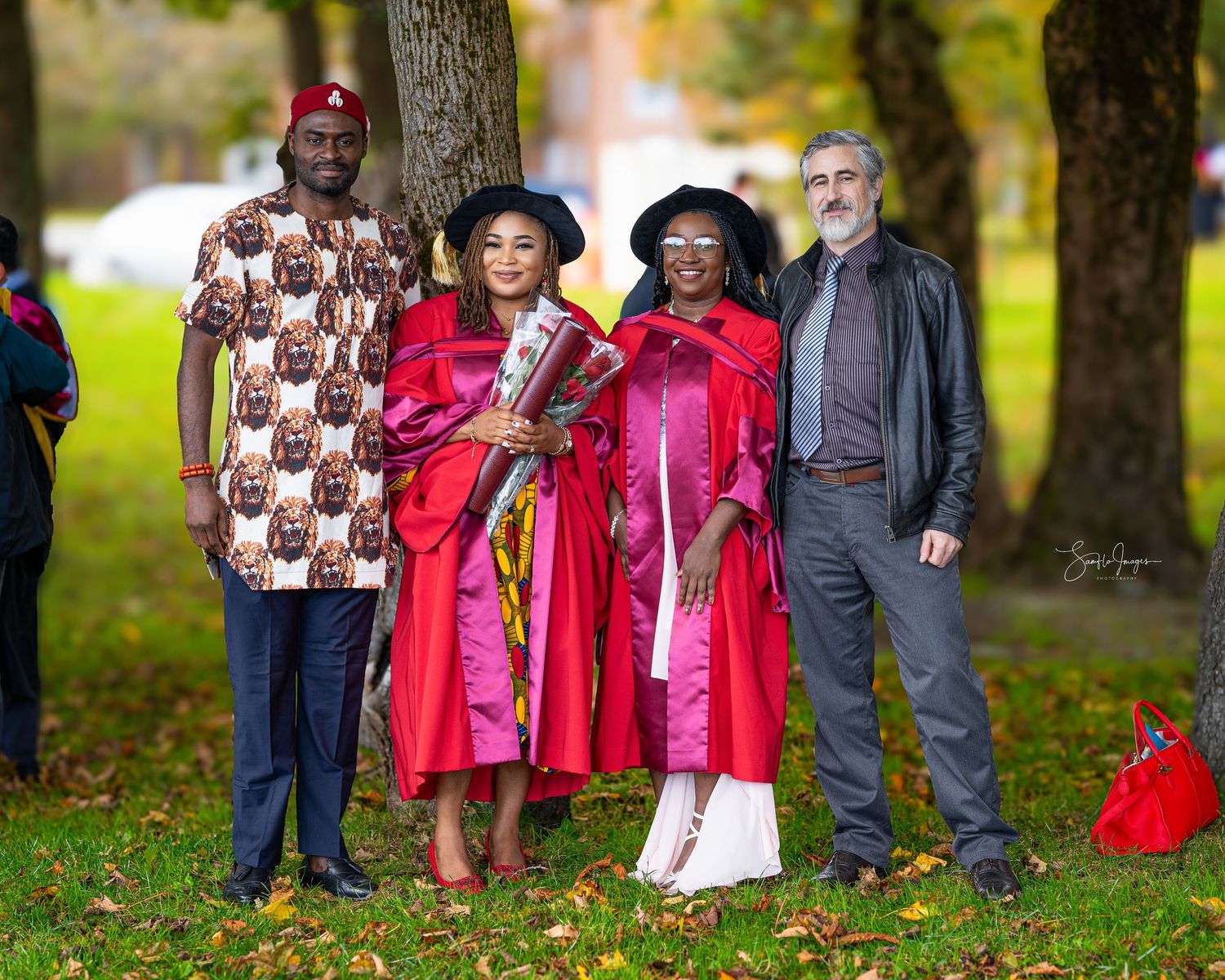 A group of people in graduation robes and formal attire stand together outdoors on an autumn day among fallen leaves.
