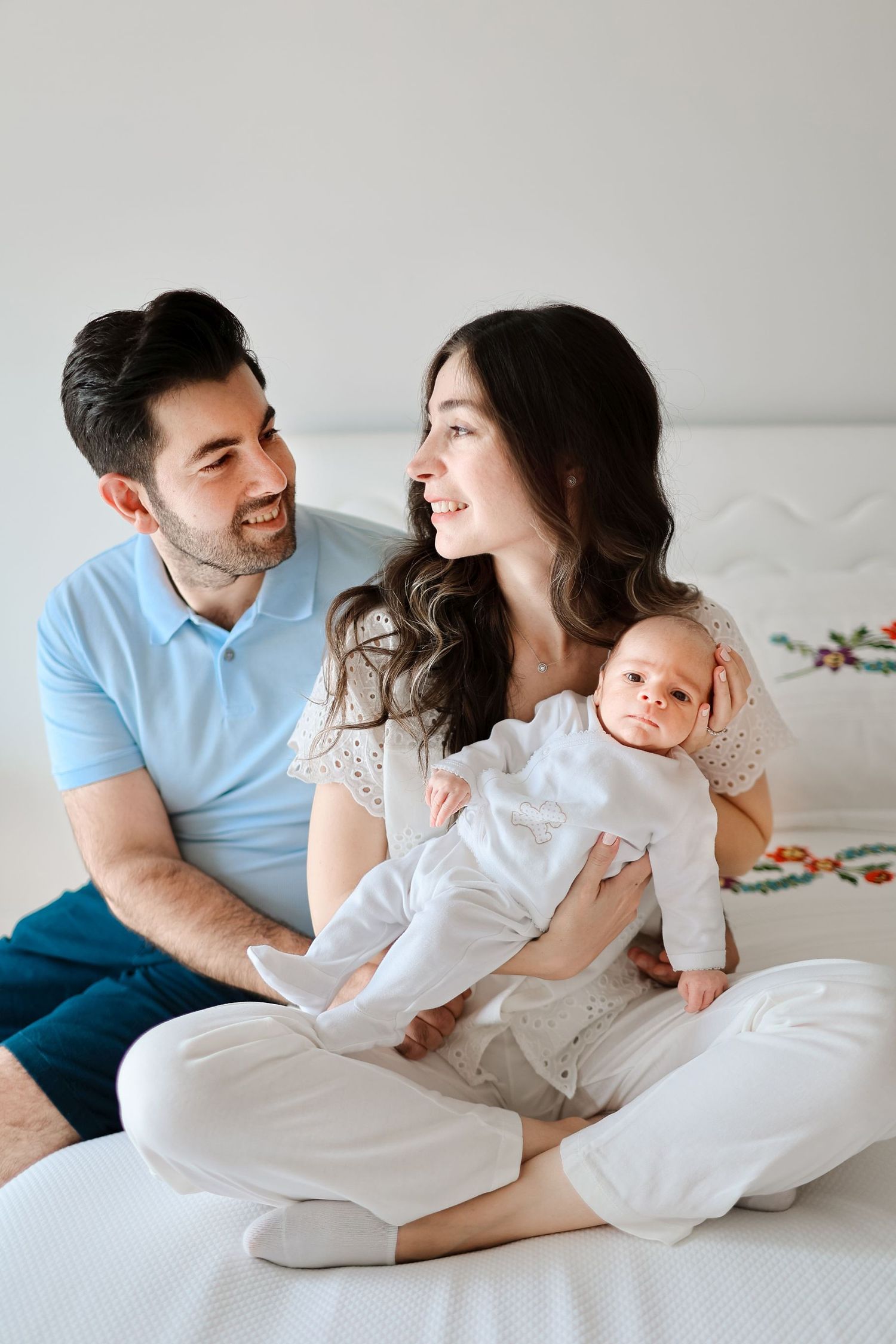 A family relaxes together on a white bed with their newborn baby in a bright, minimalist setting.