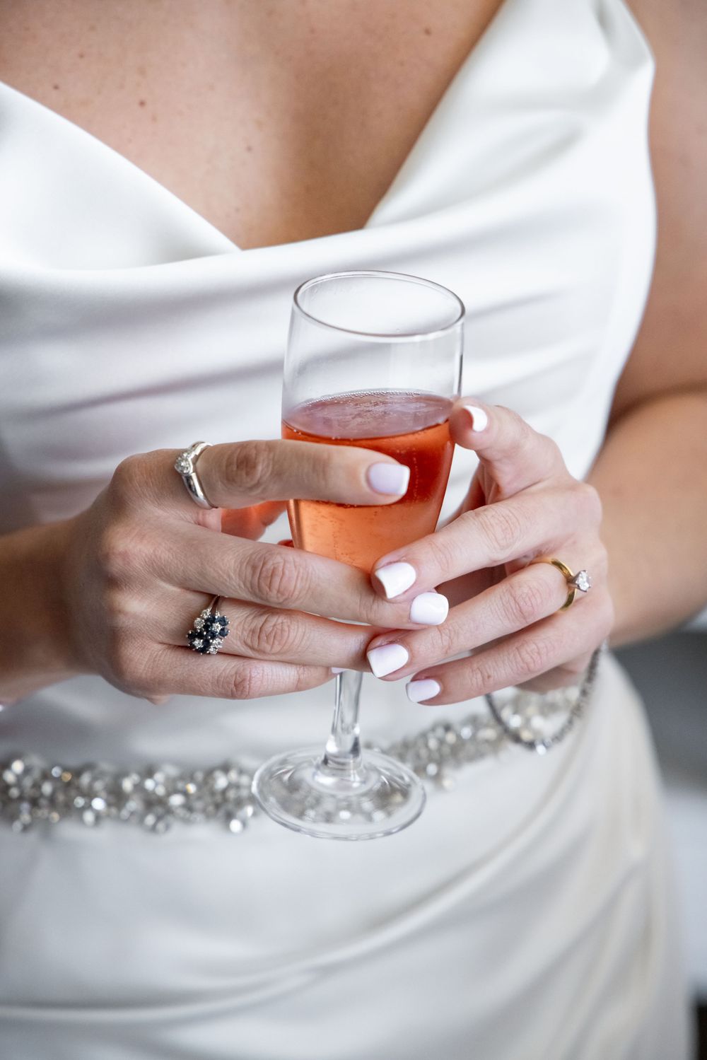 Elegant close-up of hands holding a champagne glass, showing white manicure and wedding jewelry.