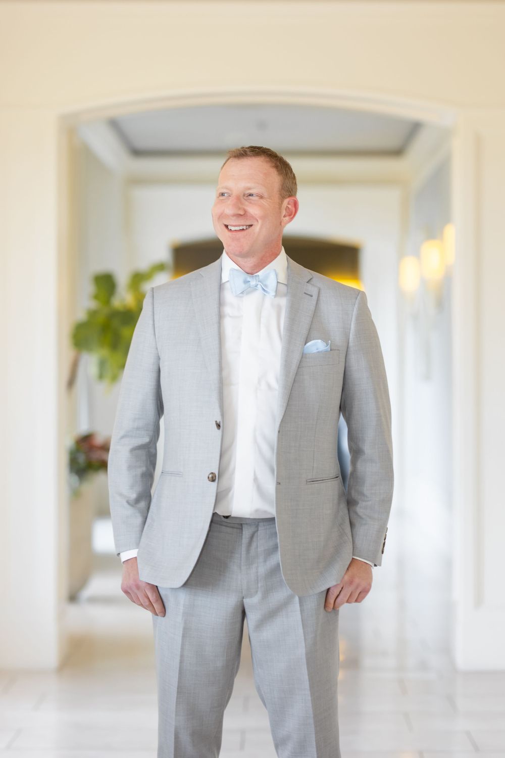 Groom wearing a light grey suit with powder blue tie stands in an elegant hallway.