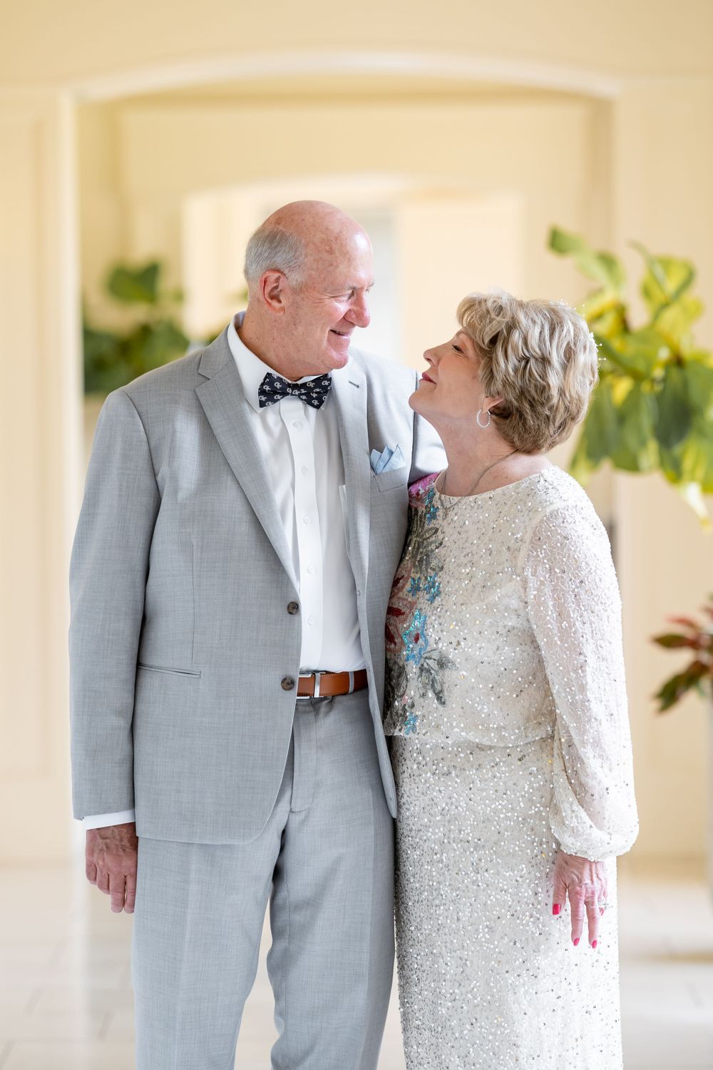 Elegant older couple in formal attire sharing a joyful moment against a cream-colored backdrop.