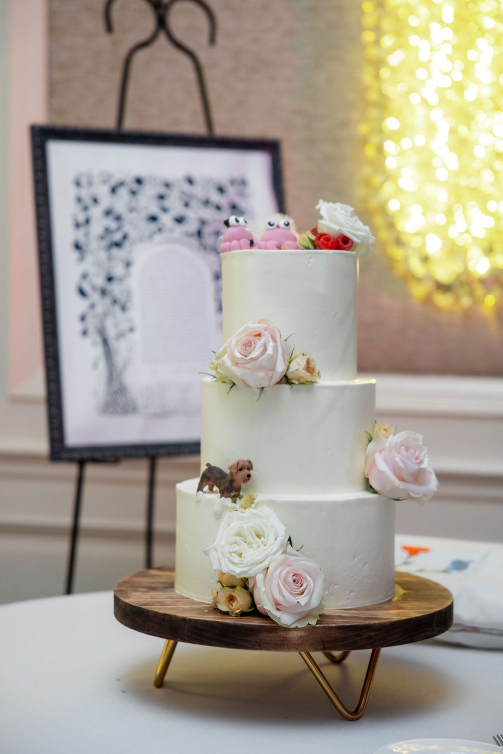 Elegant three-tier white wedding cake decorated with pink and white roses on a wooden stand.