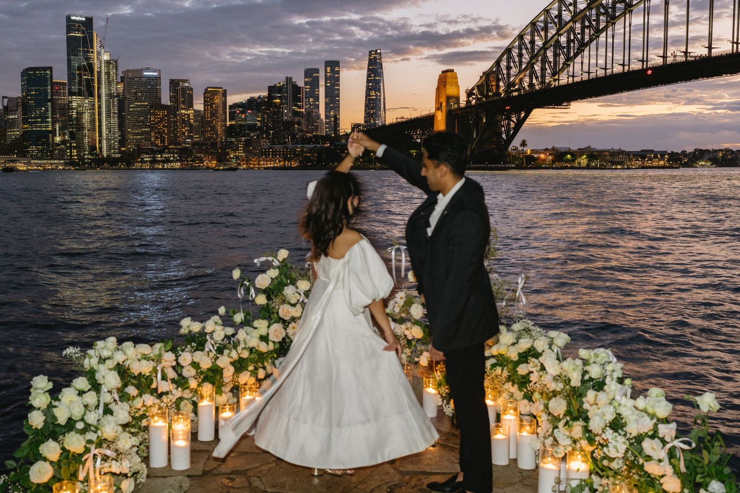 Romantic couple shares an intimate moment at sunset with Sydney Harbour Bridge and Opera House in the background.