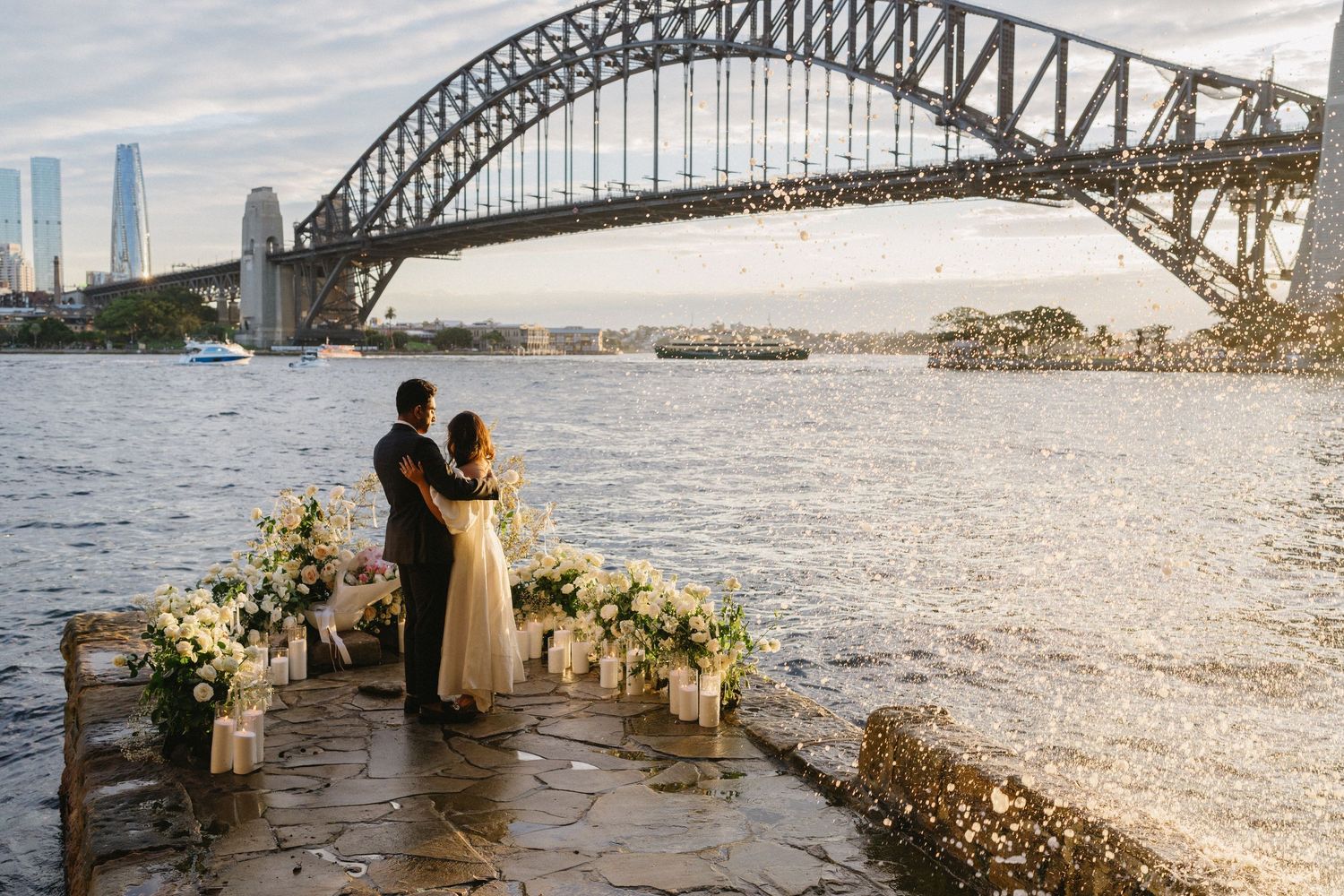Romantic couple stands silhouetted against Sydney Harbour Bridge during sunset with floral arrangements along waterfront.
