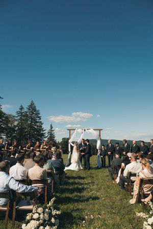 Outdoor wedding ceremony on a green hillside with guests seated along an aisle leading to a wooden arch draped in white fabric.