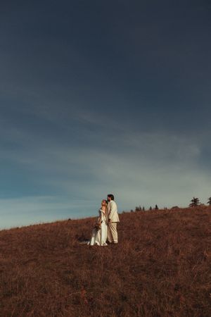 A couple in white wedding attire stands on a brown hillside against a moody cloudy sky at sunset.