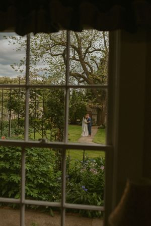 View through a window pane of a garden scene at dusk with two figures embracing on a path among trees and shrubs.