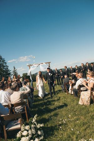 Bride and groom walk down outdoor aisle after wedding ceremony on sunny day with guests cheering on green hillside.