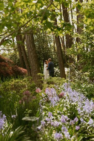 Two figures stand together in a lush garden setting with purple iris flowers blooming in the foreground and tall trees above.