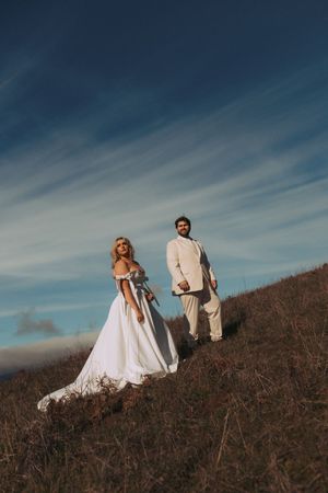 A couple in wedding attire stands on a hillside against a dramatic blue sky with wispy clouds.
