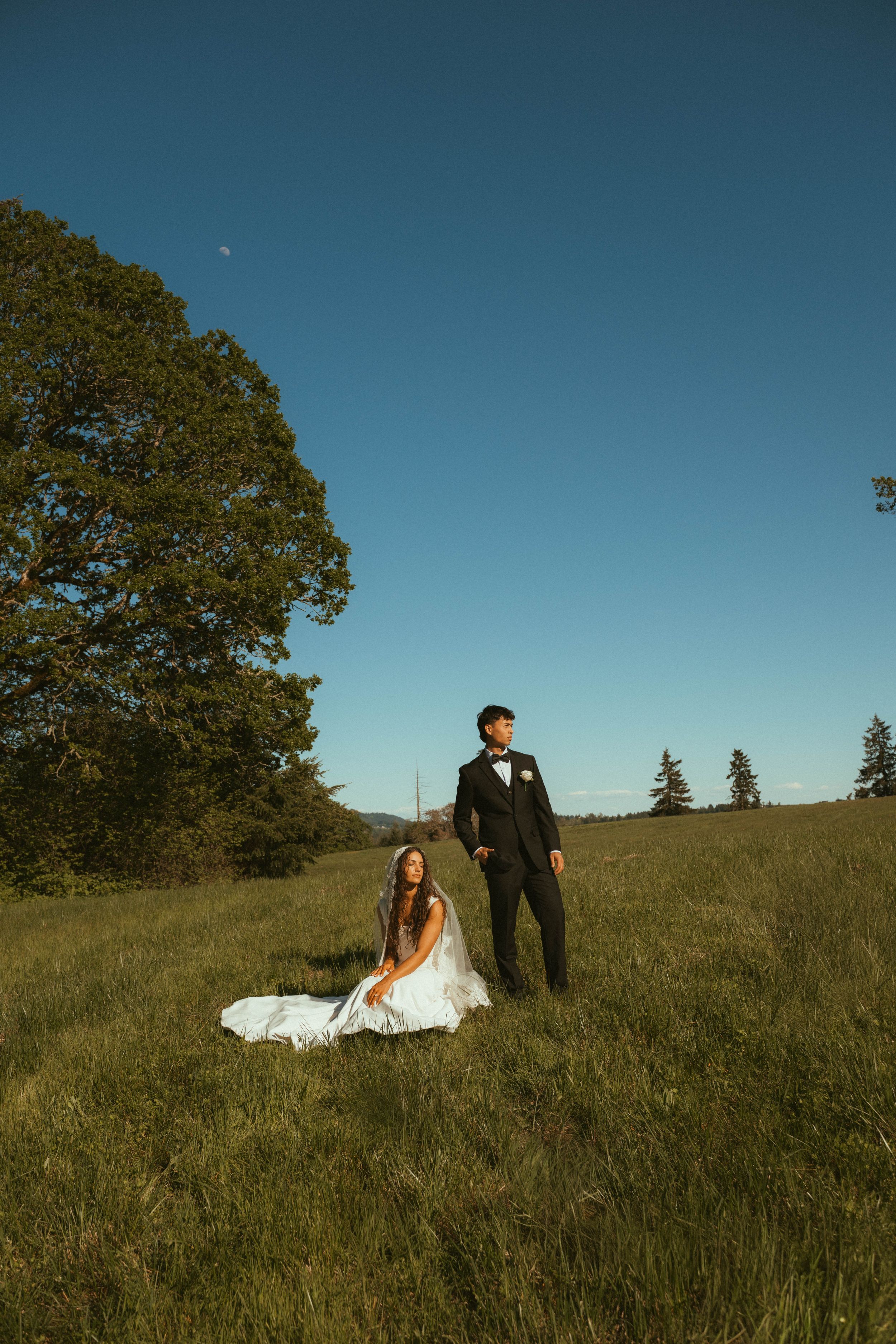 Bride in white gown sits in green meadow while groom in black suit stands beside her under clear blue sky.
