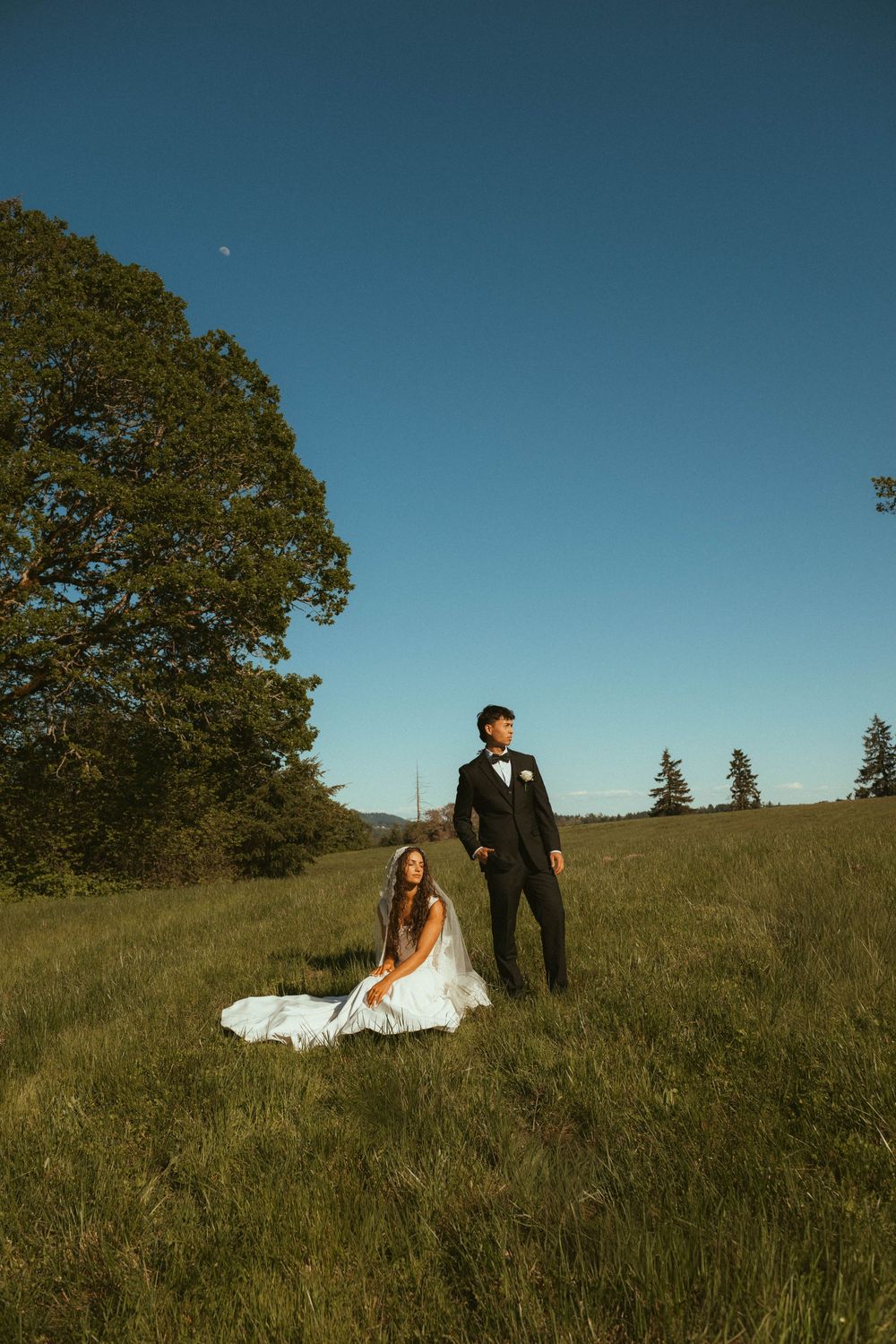 Bride in white gown sits in green meadow while groom in black suit stands beside her under clear blue sky.