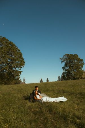 Bride and groom sit together in a sunlit green field surrounded by trees under a clear blue sky.