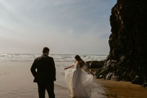 Couple on rocky beach with flowing white dress walking along ocean shore at sunset.