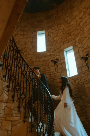 A couple in wedding attire ascending a spiral stone staircase with natural light streaming through windows.