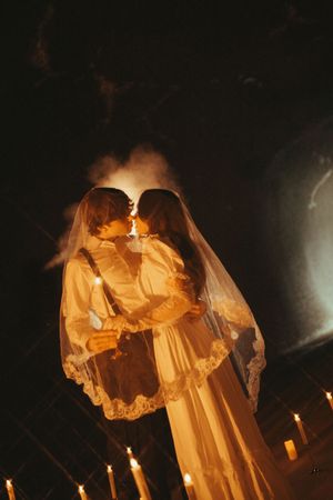 Ethereal figures in flowing white dresses hold candles in a dark atmospheric setting creating a ghostly tableau.