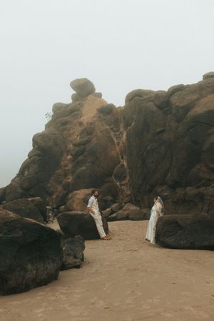 A panoramic sequence showing figures in white dresses walking between large rocky formations on a beach at dusk.