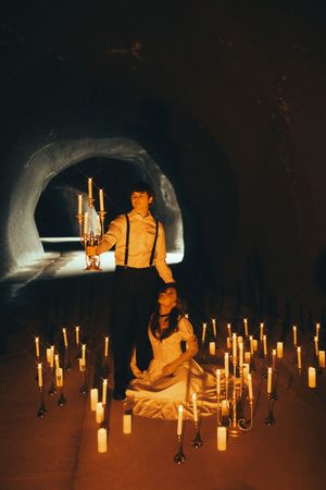 Two people surrounded by glowing candles in a dark tunnel with dramatic lighting and shadows.