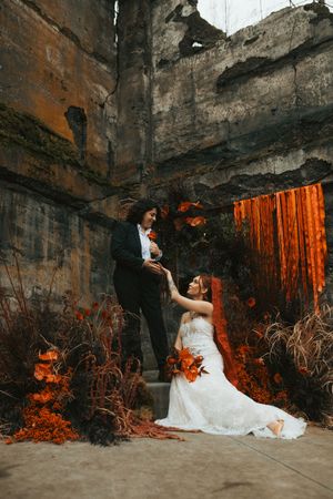 A couple poses dramatically against dark stone walls with vibrant orange autumn foliage creating a moody wedding scene.