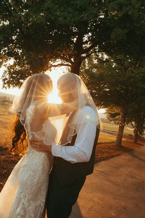 A couple in wedding attire embraces during sunset with sunlight streaming through trees in a romantic outdoor setting.