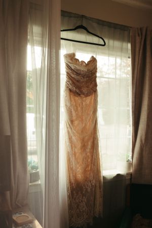 An elegant ivory lace wedding dress hangs in front of a sunlit window with sheer curtains.