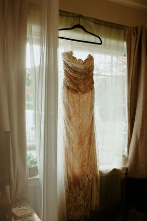 A delicate lace wedding dress hangs in a sunlit window, showcasing its elegant form and intricate details.