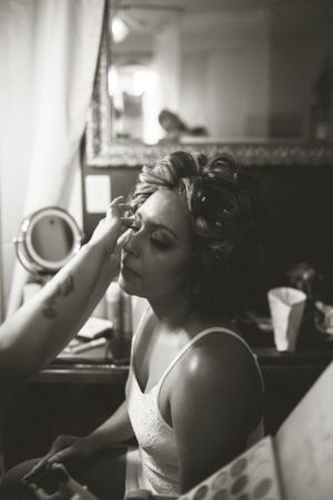 Black and white photograph of someone getting makeup done in front of a mirror, showing an intimate behind-the-scenes moment.