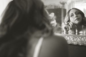 Vintage style black and white reflection in ornate mirror shows someone putting on earrings in soft lighting.