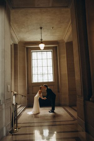 A romantic couple shares a tender moment in an elegant marble hallway with natural light streaming through a tall window.
